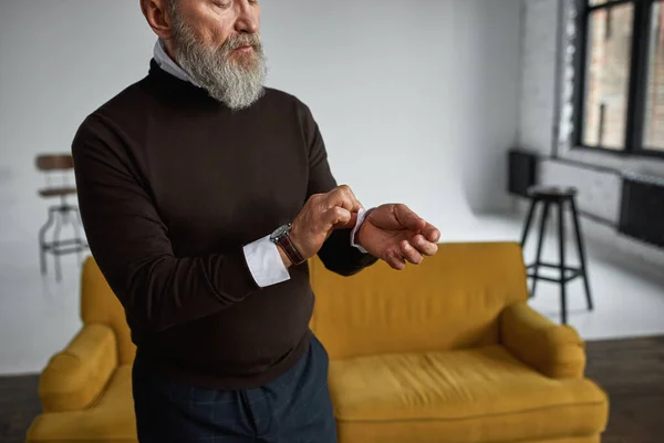 Obscure face of elderly serious caucasian man correcting shirt sleeve in spacious apartment. Trendy bearded pensioner wearing casual clothes. Concept of modern senior male lifestyle. Daytime