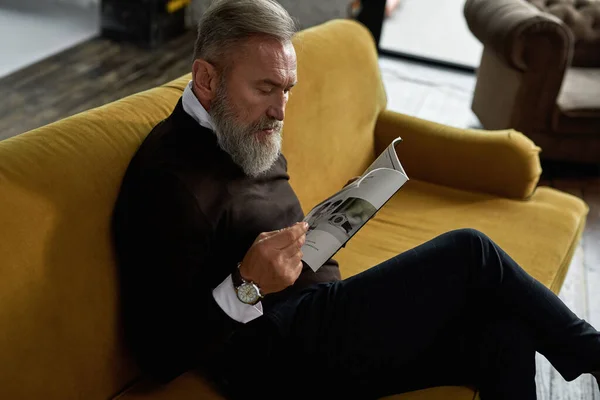 Grey hair focused caucasian man reading journal on sofa in spacious flat. Fashionable bearded pensioner wearing casual clothes. Modern senior male lifestyle. Domestic rest and leisure