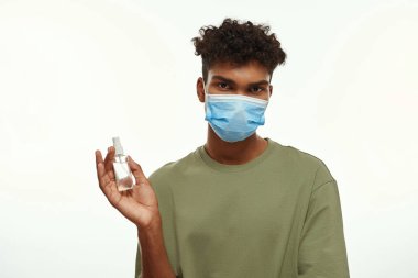 Black guy wearing medical mask holding sanitizer in hand. Partial young brunette curly man. Health protection during Coronavirus pandemic. Isolated on white background. Studio shoot. Copy space