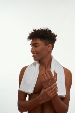 Cropped image of black guy smearing cosmetic on his hand. Young smiling brunette curly man with towel and naked torso. Body and skin care. Isolated on white background. Studio shoot. Copy space
