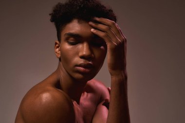 Cropped image of black beautiful guy posing with closed eyes. Young brunette curly man with naked torso. Concept of modern male beauty. Isolated on brown background. Studio shoot. Copy space