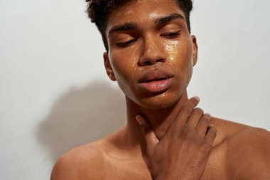 Partial of concentrated black guy with golden mask on his face. Young brunette curly man with clean face and naked torso. Facial skin care. Isolated on white background. Studio shoot
