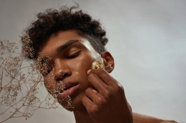 Cropped of black guy applying ice slice with plant inside on his face. Young man with closed eyes. Body and skin care. Concept of natural cosmetic. White brown background. Studio shoot. Copy space