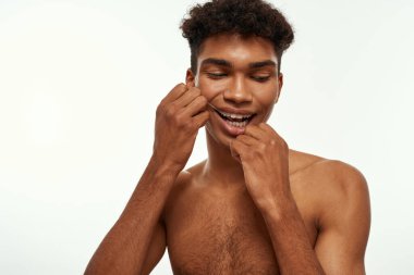 Partial of black guy brushing his teeth with dental floss. Young brunette curly man with naked torso. Dental care and hygiene. Isolated on white background. Studio shoot. Copy space