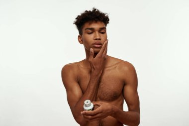 Serious black guy smearing face with after shave gel. Young slim brunette curly man with naked torso. Hygiene and skin care. Isolated on white background. Studio shoot. Copy space