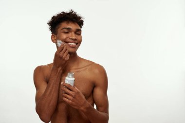 Smiling black guy with closed eyes hold jar with after shave gel and apply feather to face. Young man with naked torso and clean face. Body and skin care. White background. Studio shoot. Copy space