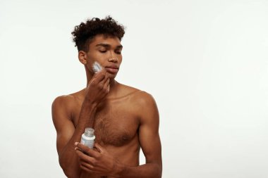 Black guy holding jar with after shave gel and applying feather to his face. Young man with naked torso and clean face. Body and skin care. Isolated on white background. Studio shoot. Copy space