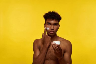 Confident black guy smearing cosmetic cream from container on his face. Young man with naked torso looking at camera. Facial skin care. Isolated on yellow background. Studio shoot. Copy space