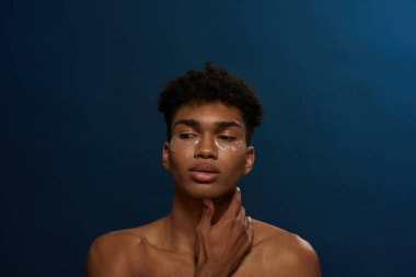 Cropped of thoughtful black guy with under eye patches on face. Young brunette curly man. Body and skin care. Facial lifting and rejuvenation. Isolated on blue background. Studio shoot. Copy space