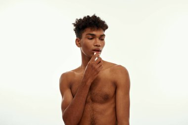 Partial image of black guy moisturizing his lips with hygienic lipstick. Young brunette curly man with naked torso. Body and skin care. Isolated on white background. Studio shoot. Copy space