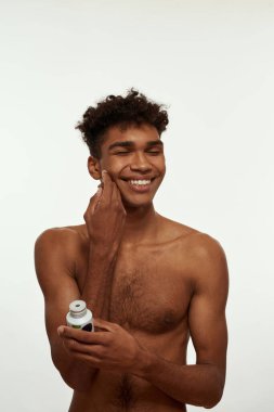 Smiling black guy with closed eyes smearing face with after shave gel. Young slim brunette curly man with naked torso. Hygiene and skin care. Isolated on white background. Studio shoot. Copy space