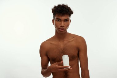 Serious black guy holding antiperspirant deodorant and looking at camera. Young brunette curly man with naked torso. Male beauty and body care. Isolated on white background. Studio shoot. Copy space
