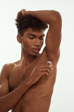 African american guy applying deodorant on his armpit. Young fit brunette curly man with naked torso. Male beauty and body care. Isolated on white background. Studio shoot. Copy space