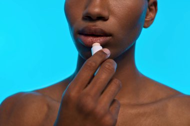 Siyahi bir adamın hijyenik rujla dudak boyamasının kısmi görüntüsü. Çıplak gövdeli, sıska bir gencin belirsiz yüzü. Vücut ve cilt bakımı. Açık mavi arka planda izole edilmiş. Stüdyo çekimi