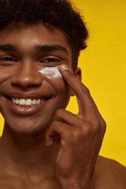Yüzüne kozmetik krem süren gülümseyen bir zenci. Kameraya bakan esmer, kıvırcık adamın belirsiz yüzü. Yüz bakımı. Sarı arka planda izole edilmiş. Stüdyo çekimi