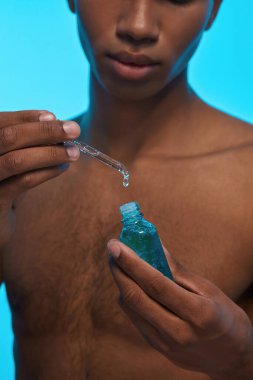 Partial of black guy holding dropper and bottle with natural organic cosmetic oil. Obscure face of young slim man with naked torso. Body and skin care. Isolated on light blue background. Studio shoot