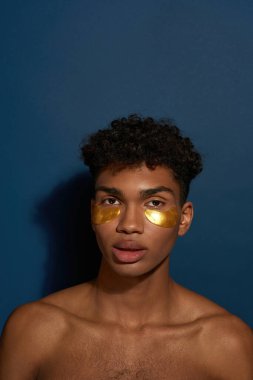Yüzünde yamalar olan ciddi siyahi bir adam. Çıplak gövdeli genç adam kameraya bak. Vücut ve cilt bakımı. Yüz gerdirme ve gençleştirme. Mavi arka plan. Stüdyo çekimi. Boşluğu kopyala