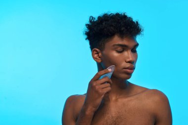 Partial image of black guy applying ice slice on his face. Young slim brunette curly man with naked torso. Body and skin care. Isolated on light blue background. Studio shoot. Copy space