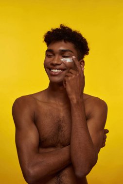 Yüzüne kozmetik krem süren gülümseyen bir zenci. Çıplak gövdeli, zayıf, esmer, kıvırcık adam başka tarafa bakıyor. Yüz bakımı. Sarı arka planda izole edilmiş. Stüdyo çekimi