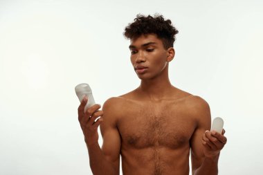 Elinde deodorant ve kağıt tutan konsantre siyahi bir adam. Çıplak gövdeli esmer kıvırcık adam. Erkek güzelliği ve vücut bakımı. Beyaz arka planda izole edilmiş. Stüdyo çekimi. Boşluğu kopyala