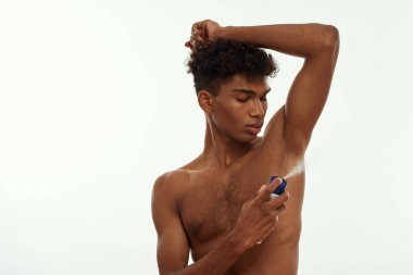 Siyah adam koltuk altına sprey deodorant sürüyor. Çıplak gövdeli, zayıf, esmer, kıvırcık adam. Erkek güzelliği ve vücut bakımı. Beyaz arka planda izole edilmiş. Stüdyo çekimi. Boşluğu kopyala