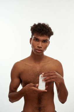 Elinde deodorant tutan ve kameraya bakan ciddi siyahi bir adamın ön görüntüsü. Çıplak gövdeli esmer kıvırcık adam. Erkek güzelliği ve vücut bakımı. Beyaz arka planda izole edilmiş. Stüdyo çekimi. Boşluğu kopyala