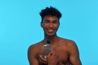 Smiling african american guy holding and pointing with finger at bottle with shower gel. Young man with naked torso. Hygiene and body care. Isolated on light blue background. Studio shoot. Copy space