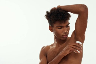 Siyah adam koltuk altına deodorant sürüyor. Çıplak gövdeli genç, ince, esmer, kıvırcık adam. Erkek güzelliği ve vücut bakımı. Beyaz arka planda izole edilmiş. Stüdyo çekimi. Boşluğu kopyala