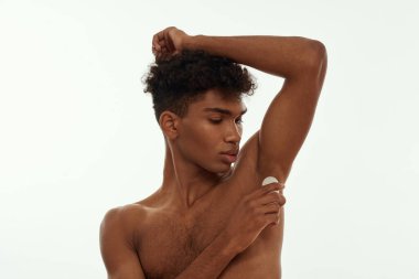 Black guy applying antiperspirant deodorant on his armpit. Young brunette curly man with naked torso. Male beauty and body care. Isolated on white background. Studio shoot. Copy space
