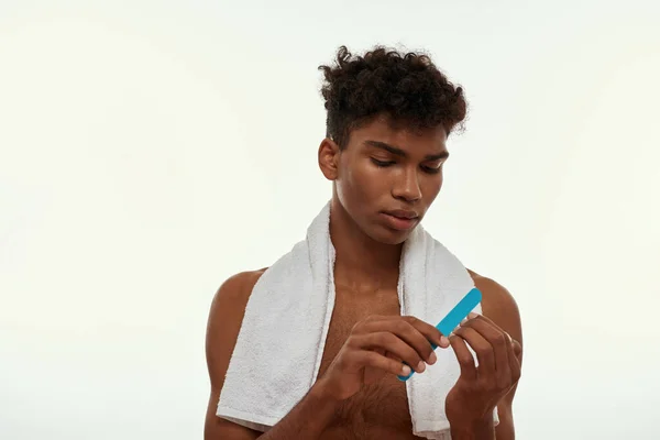 Partial image of black guy doing manicure of fingernails with nail file. Young focused brunette curly man with towel and naked torso. Body care and hygiene. White background. Studio shoot. Copy space
