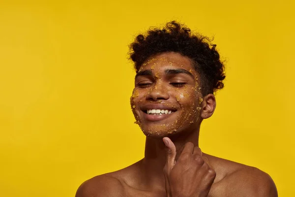 Partial of smiling black guy with honey scrub on his face skin. Young brunette curly man with closed eyes. Facial skin care. Isolated on yellow background. Studio shoot. Copy space