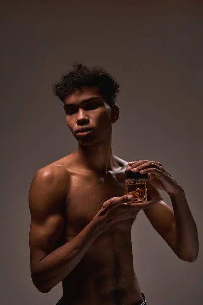 Handsome fit black guy holding luxury aromatic perfume. Young brunette curly man with naked torso. Concept of modern male beauty. Isolated on brown background. Studio shoot. Copy space