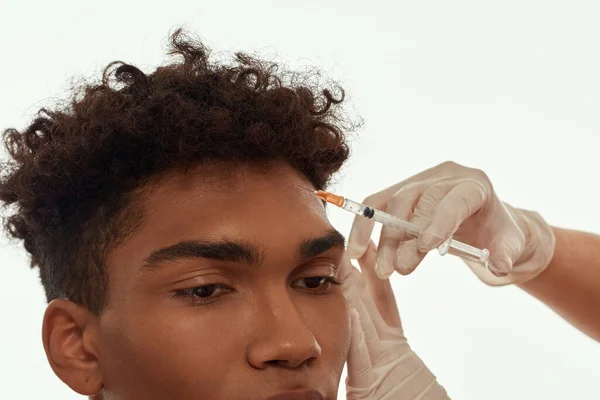 Doctor hands wearing latex gloves making injection of botox with syringe on face of black guy. Obscure face of y young curly man. Face lifting and mesotherapy. Isolated on white background in studio