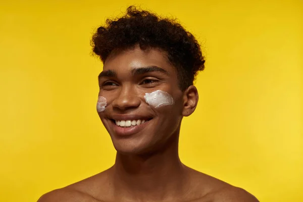 Partial image of smiling black guy with cosmetic cream on his face looking away. Young brunette curly man. Facial skin care. Isolated on yellow background. Studio shoot. Copy space