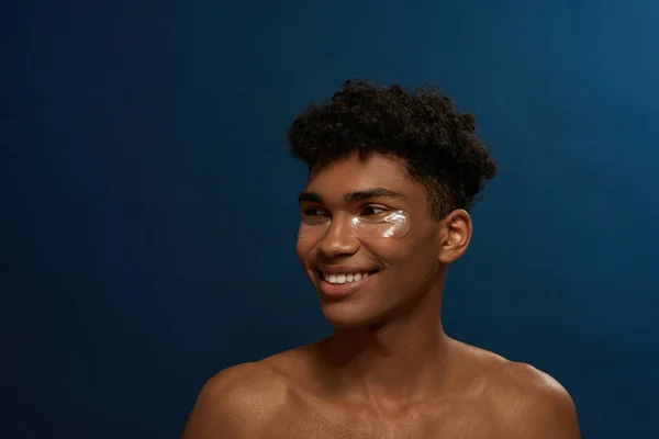 Cropped of smiling black guy with under eye patches on face look away. Young brunette curly man. Body and skin care. Facial lifting and rejuvenation. Isolated on blue background in studio. Copy space
