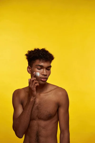 African american guy smearing cosmetic cream on his face. Young brunette curly man with naked torso. Concept of facial skin care. Isolated on yellow background. Studio shoot. Copy space