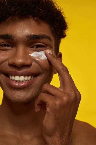 Partial of smiling black guy smearing cosmetic cream on his face. Obscure face of young brunette curly man looking at camera. Facial skin care. Isolated on yellow background. Studio shoot