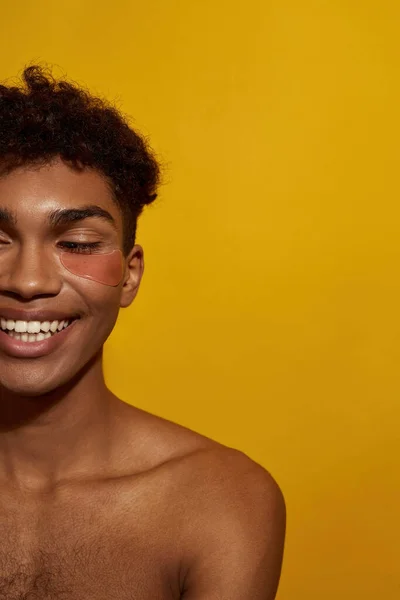 Cropped of smiling black guy with under eye patch on face. Obscure face of young man with naked torso. Body and skin care. Facial lifting and rejuvenation. Yellow background. Studio shoot. Copy space