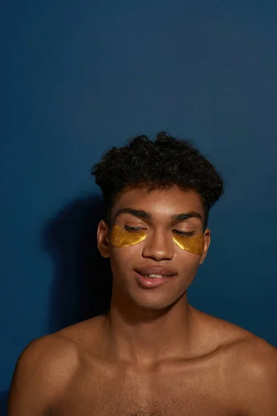 Partial of cunning black guy with under eye patches on face looking away. Young man with naked torso. Body and skin care. Facial lifting and rejuvenation. Blue background. Studio shoot. Copy space