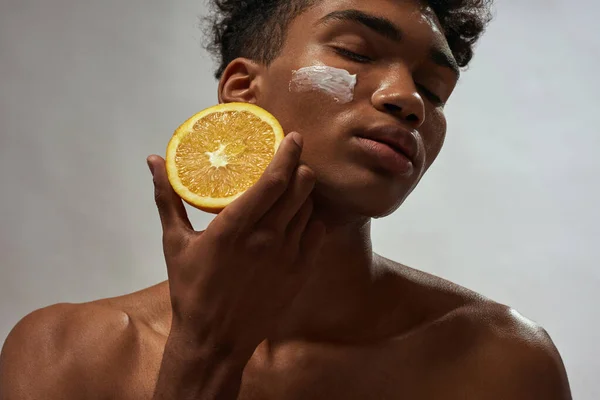 Cropped of black guy holding orange with cosmetic cream on his face on this fruit. Young brunette curly man with closed eyes. Facial skin care. Isolated on white brown background. Studio shoot