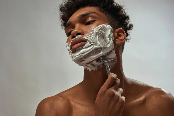 Cropped of black guy shaving razor with shaving foam. Young brunette curly man with naked torso. Hygiene and skin care. Isolated on white brown background. Studio shoot. Copy space
