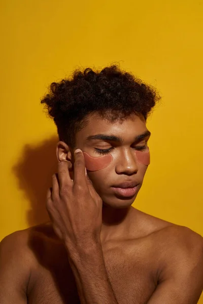 Cropped image of black guy applying or correcting under eye patch on face. Young brunette curly man. Body and skin care. Facial lifting and rejuvenation. Yellow background. Studio shoot. Copy space
