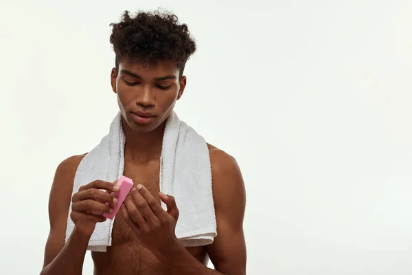 Partial image of black guy doing manicure of fingernails with nail buffer block. Young brunette curly man with towel and naked torso. Body care and hygiene. White background. Studio shoot. Copy space