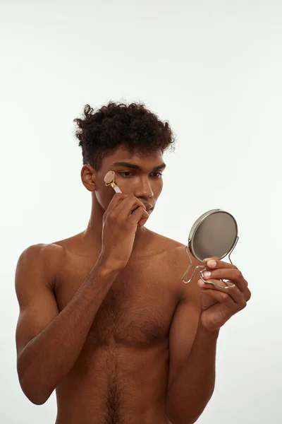 Cropped image of black guy looking at mirror massaging his face with jade roller. Young focused curly man with naked torso. Body and skin care. Isolated on white background. Studio shoot. Copy space