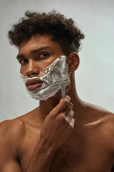 Cropped of black guy shaving razor with shaving foam. Young brunette curly man with naked torso looking at camera. Hygiene and skin care. Isolated on white brown background. Studio shoot