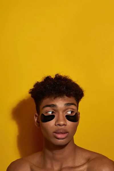 Cropped of focused black guy with under eye patches on face looking away. Young brunette curly man. Body and skin care. Facial lifting and rejuvenation. Yellow background. Studio shoot. Copy space