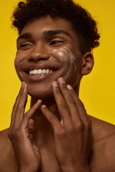 Partial of smiling black guy smearing cosmetic cream on his face. Young brunette curly man with clean face looking away. Facial skin care. Isolated on yellow background. Studio shoot