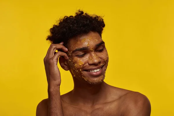 Cropped of smiling black guy with honey scrub on his face skin. Young brunette curly man with closed eyes. Facial skin care. Isolated on yellow background. Studio shoot. Copy space