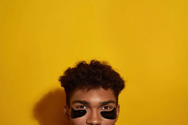 Obscure face of black guy with under eye patches on face. Young brunette curly man looking at camera. Body and skin care. Facial lifting and rejuvenation. Yellow background. Studio shoot. Copy space