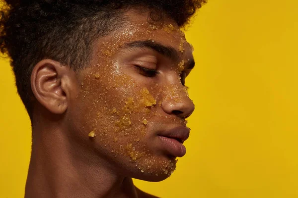 Profile of black guy with honey scrub on his face skin. Cropped of young brunette curly man with closed eyes. Concept of facial skin care. Isolated on yellow background. Studio shoot